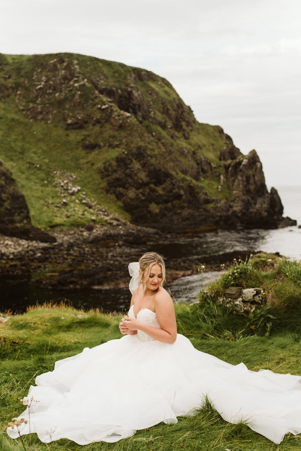Bride sitting on the grass at Kinbane Castle in Ireland wearing a strapless lace and tulle ballgown and a tulle bow in her hair.