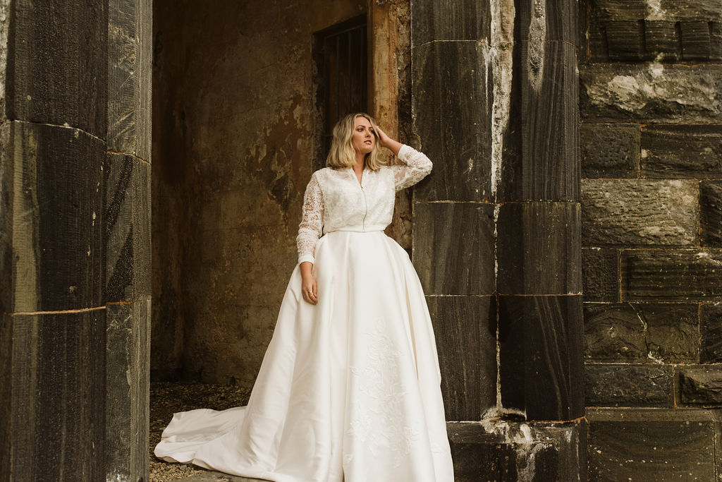 Bride standing in doorway of abandoned church wearing a mikado ballgown wedding dress and lace bomber jacket at Dunlewey Church in Ireland