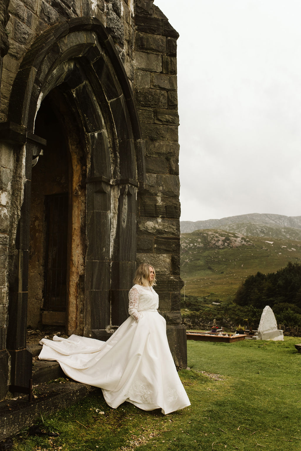 Bride walking out of the doorway of abandoned church with her hands in the pockets of her mikado ballgown wedding dress and lace bomber jacket at Dunlewey Church in Ireland