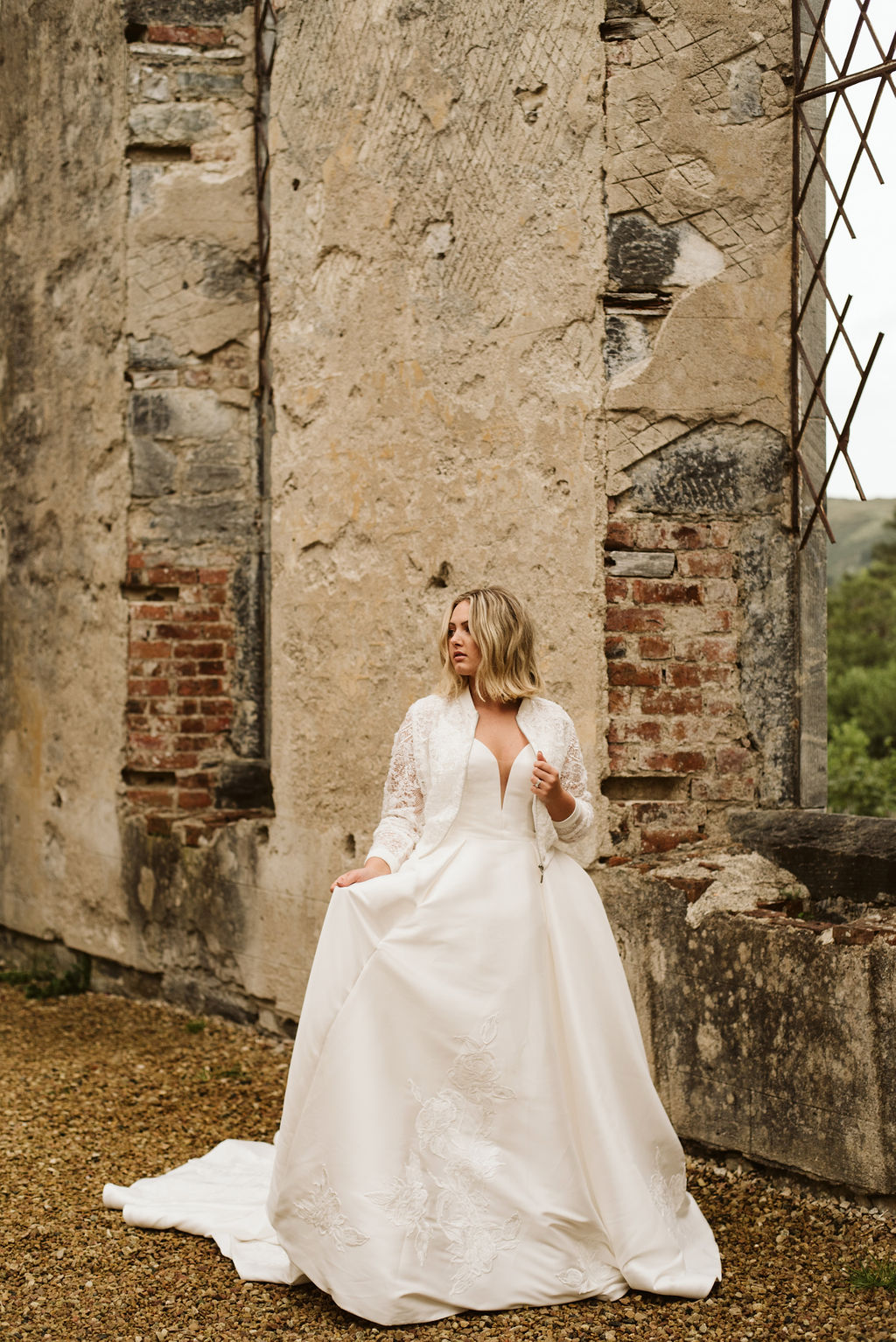 Bride walking through abandoned church wearing a mikado ballgown wedding dress with lace detail at the bottom of the skirt and a lace bomber jacket at Dunlewey Church in Ireland