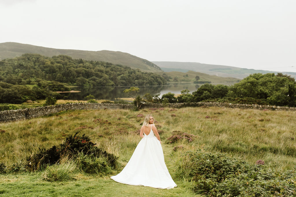 Back view of bride wearing a mikado ballgown wedding dress with straps in a field of grasses and purple flowers next to a lake at Dunlewey Church in Ireland