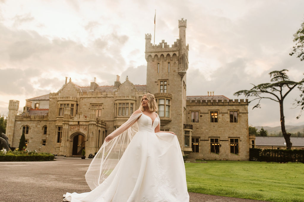 Bride wearing a mikado ballgown wedding dress with straps and tulle cape in front of Lough Eske castle in Donegal, Ireland.