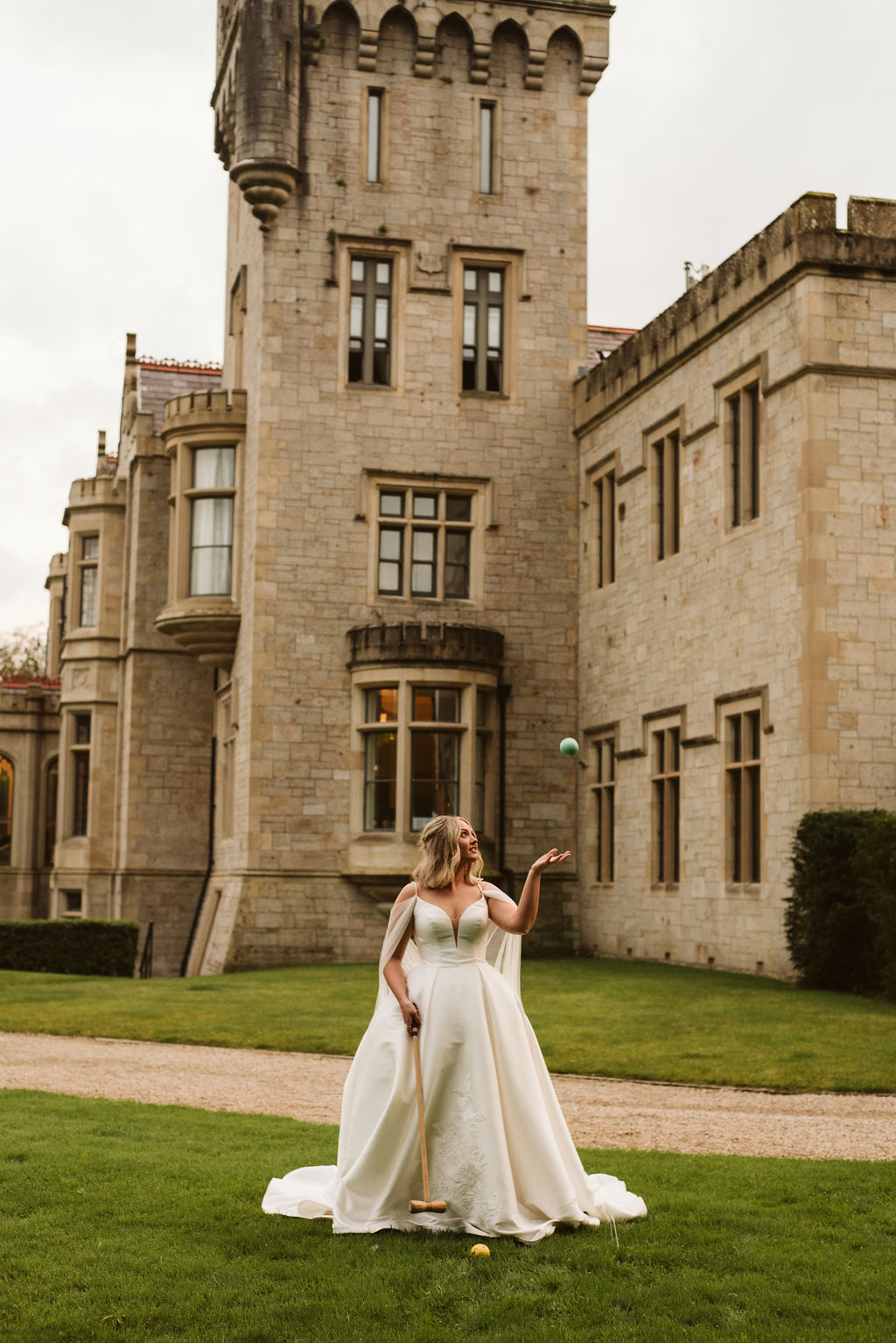 Bride wearing a mikado ballgown wedding dress with straps and tulle cape playing croquet in front of Lough Eske castle in Donegal, Ireland.