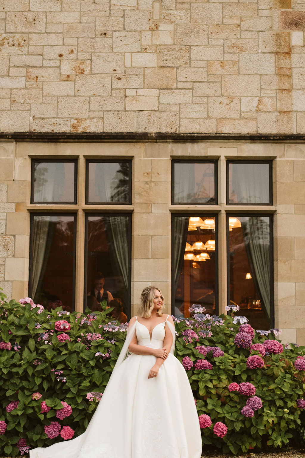 Bride wearing a mikado ballgown wedding dress with straps and tulle cape in front of hydrangeas at Lough Eske castle in Donegal, Ireland.