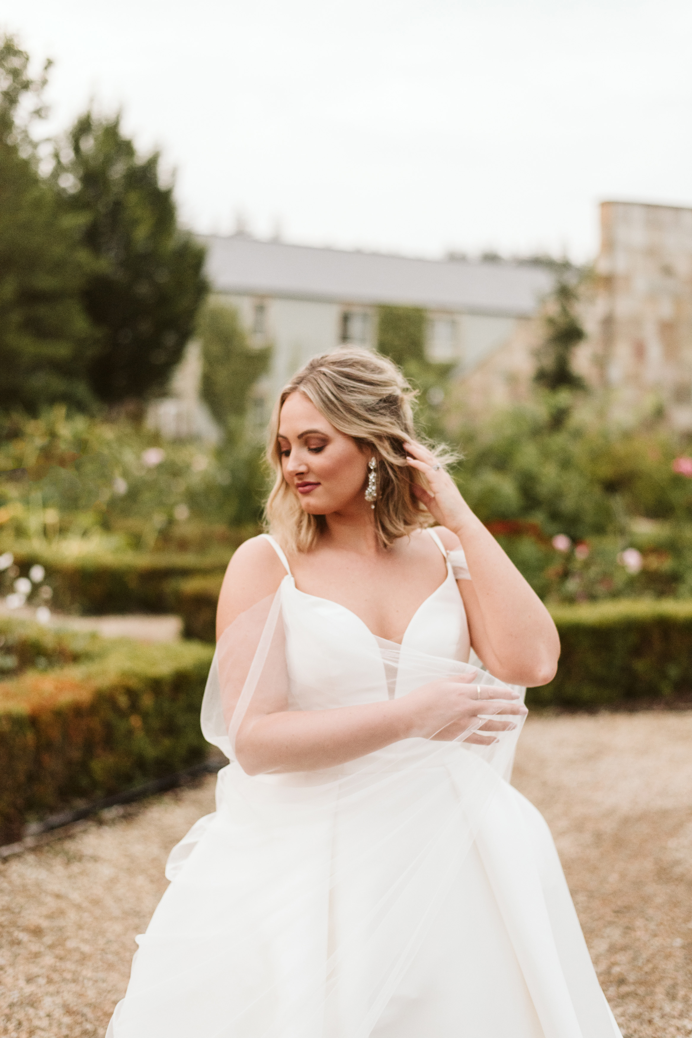 Brideshowing off her sparkly earrings in a mikado ballgown wedding dress and tulle cape at Lough Eske Castle's grounds in Ireland
