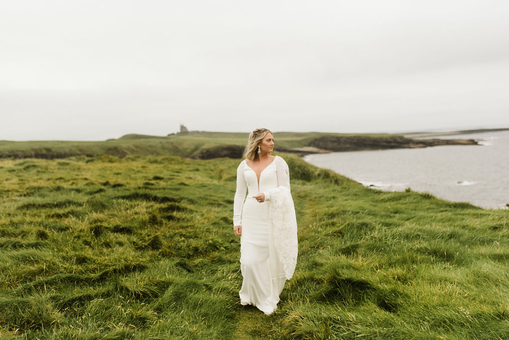 Bride carries the train of her simple fitted wedding dress with plunge neckline and long sleeves at Mullaghmore Head in Ireland