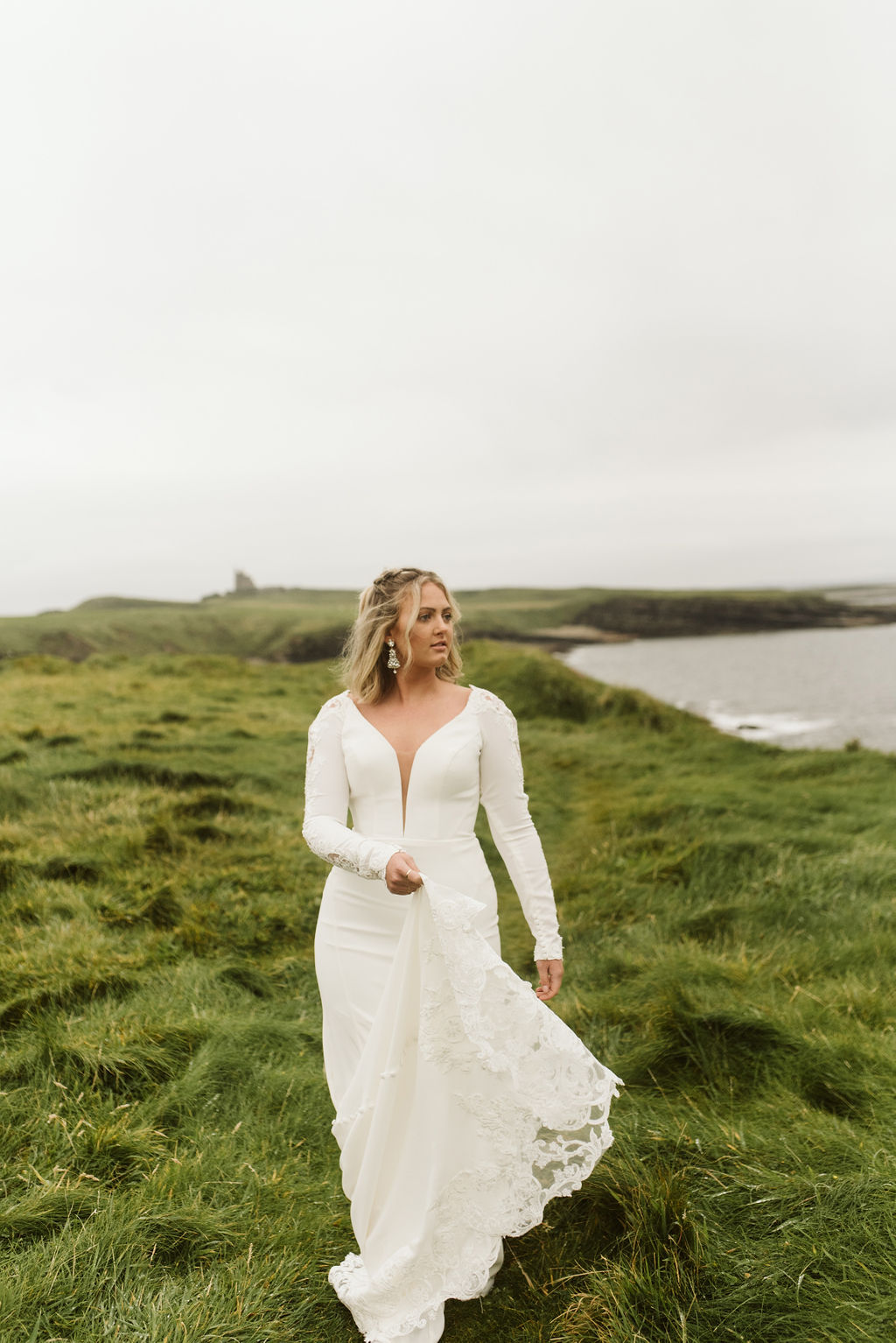Bride swings the train of her simple fitted wedding dress with plunge neckline and long sleeves at Mullaghmore Head in Ireland