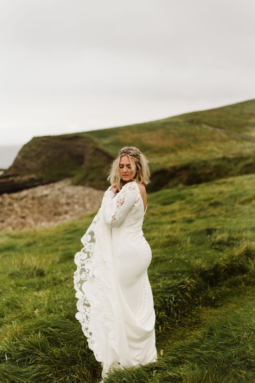 Back view of bride looking over her shoulder in a crepe fitted wedding dress with lace train and long sleeves at Mullaghmore Head in Ireland