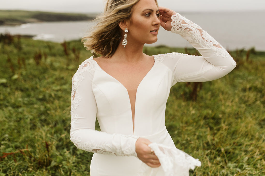 Bride wears a simple fitted wedding dress with plunge neckline and long sleeves with lace detail while looking at the Atlantic Ocean in Mullaghmore Head in Ireland