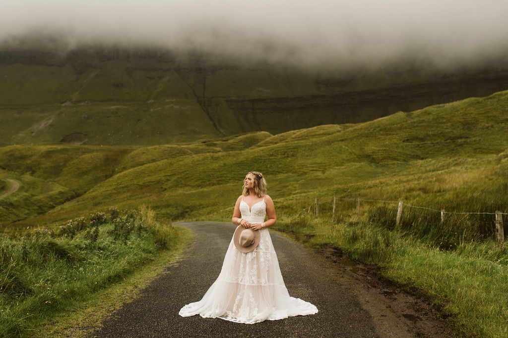 Bride in middle of road at Gleniff Horseshoe Drive in Ireland wearing a lace boho wedding dress with straps holding a rancher's hat in her hands