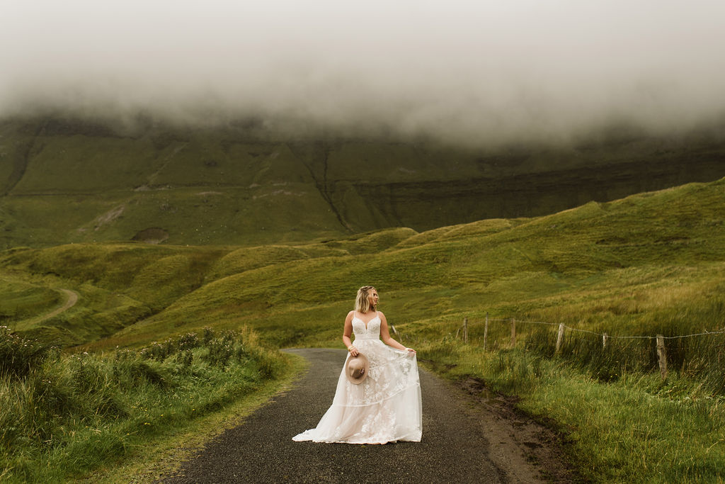Bride playing with the skirt of her lace boho wedding dress with straps while holding a rancher's hat at Gleniff Horseshoe Drive in Ireland
