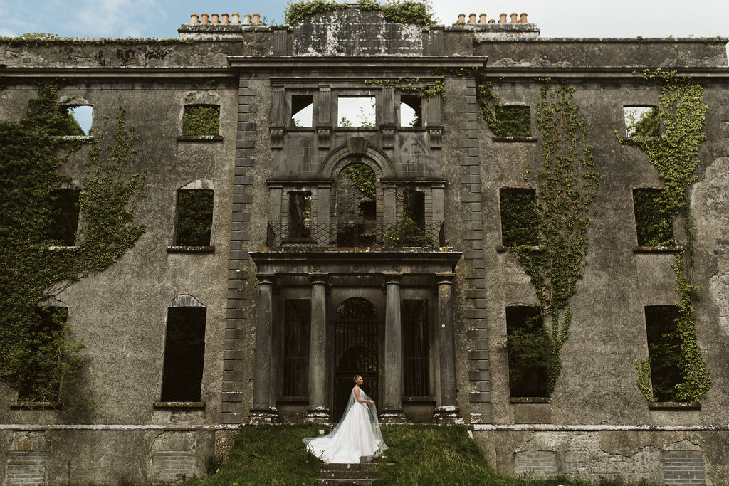 Brides stands on the steps of Moore Hall in Ireland wearing a simple ballgown wedding dress with tulle cape