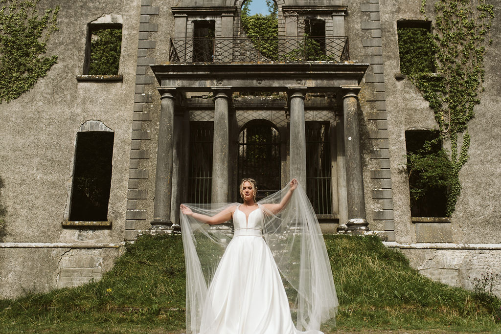 Bride plays with tulle cape in front of Moore Hall in Ireland wearing a simple ballgown wedding dress with plunging neckline