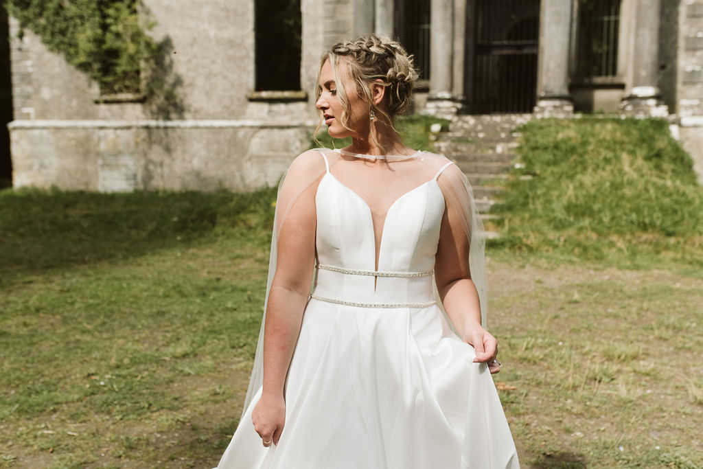 Bride with dutch braids stands in front of Moore Hall in Ireland wearing a simple ballgown wedding dress with plunging neckline, beaded waist detail and tulle bridal cape