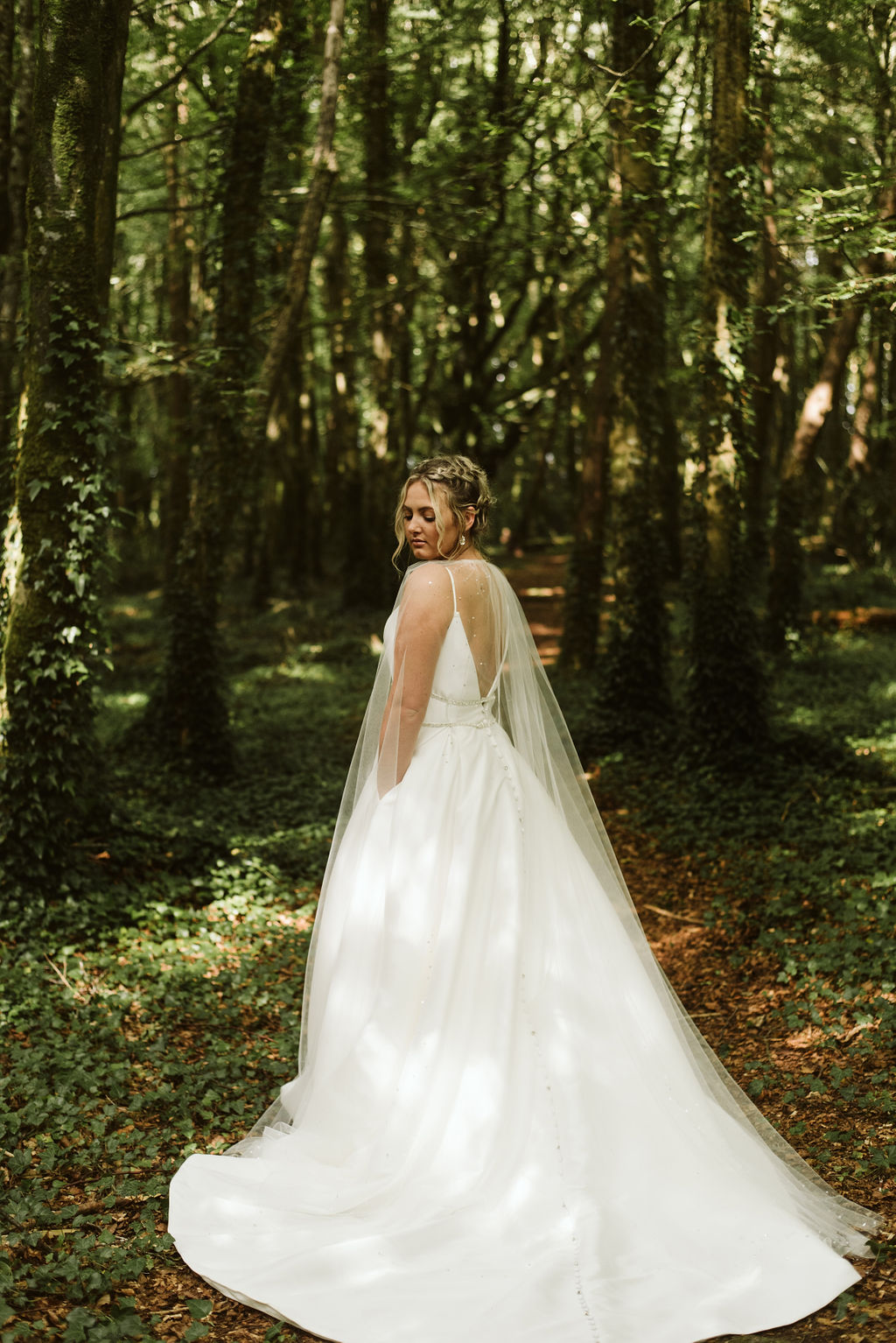 Back view of bride looking over her shoulder on the grounds of Moore Hall in Irealnd wearing a simple ballgown with open back and beaded waist detail and tulle bridal cape