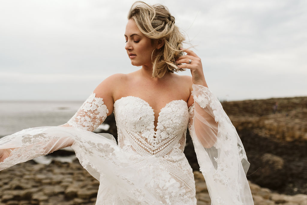 Bride plays with the lace bell sleeves of her fitted lace boho wedding dress at Giant's Causeway in Northern Ireland