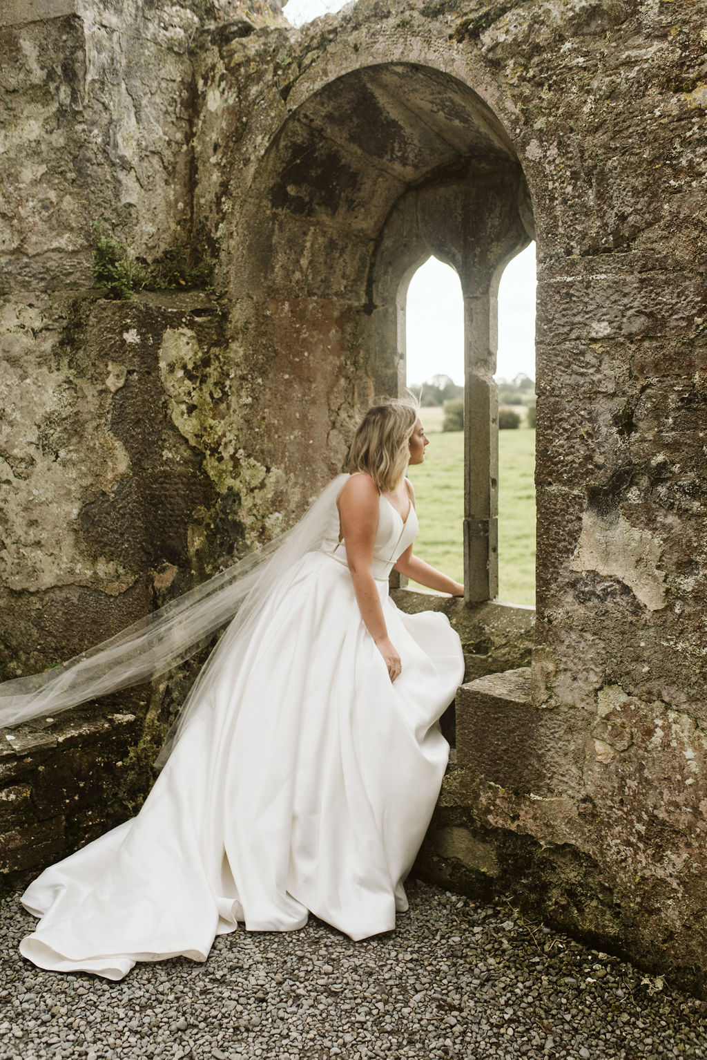 Bride looking out window of abandoned friary wearing a mikado ballgown wedding dress with beaded waist banding and tulle wings at Ross Errilly Friary in Ireland