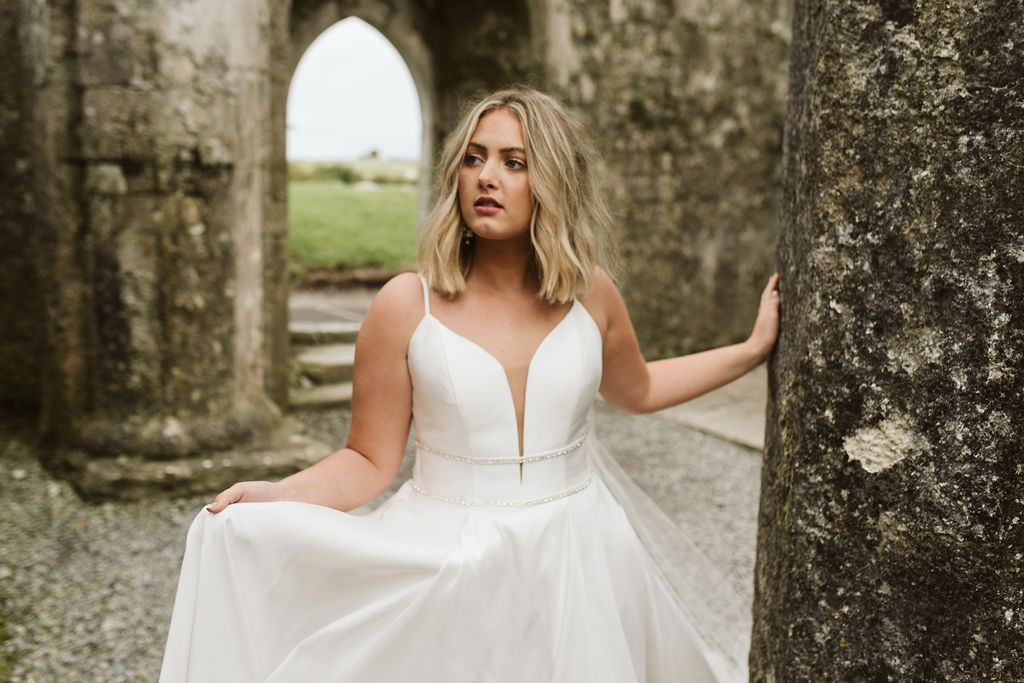 Bride wearing a mikado ballgown wedding dress with straps a plunge neckline and beaded banding at the waist walking at Ross Errilly Friary in Ireland