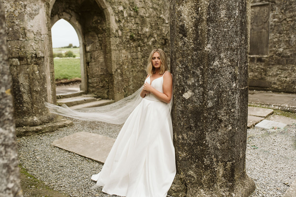 Bride leaning against column at Ross Errilly Friary wearing a mikado ballgown wedding dress with straps and tulle wings