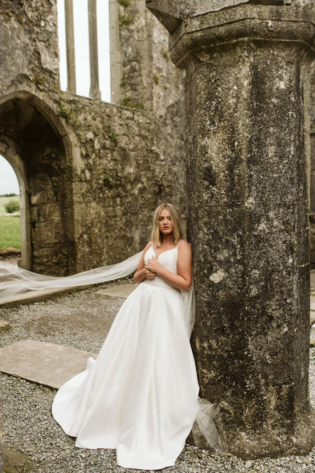 Bride standing in abadoned friary wearing a mikado ballgown wedding dress with straps and beaded waist banding with tulle wings at Ross Errilly Friary in Ireland