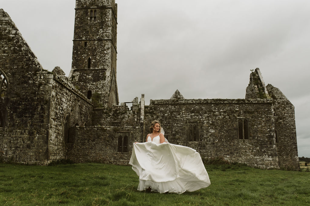 Bride wearing a mikado ballgown wedding dress with beaded waist banding runs in the field outside Ross Errilly Friary in Ireland