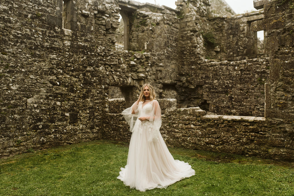 Bride wearing a lace and tulle aline wedding dress with lace straps and a tulle cape sways in the grass at Ross Errilly Friary