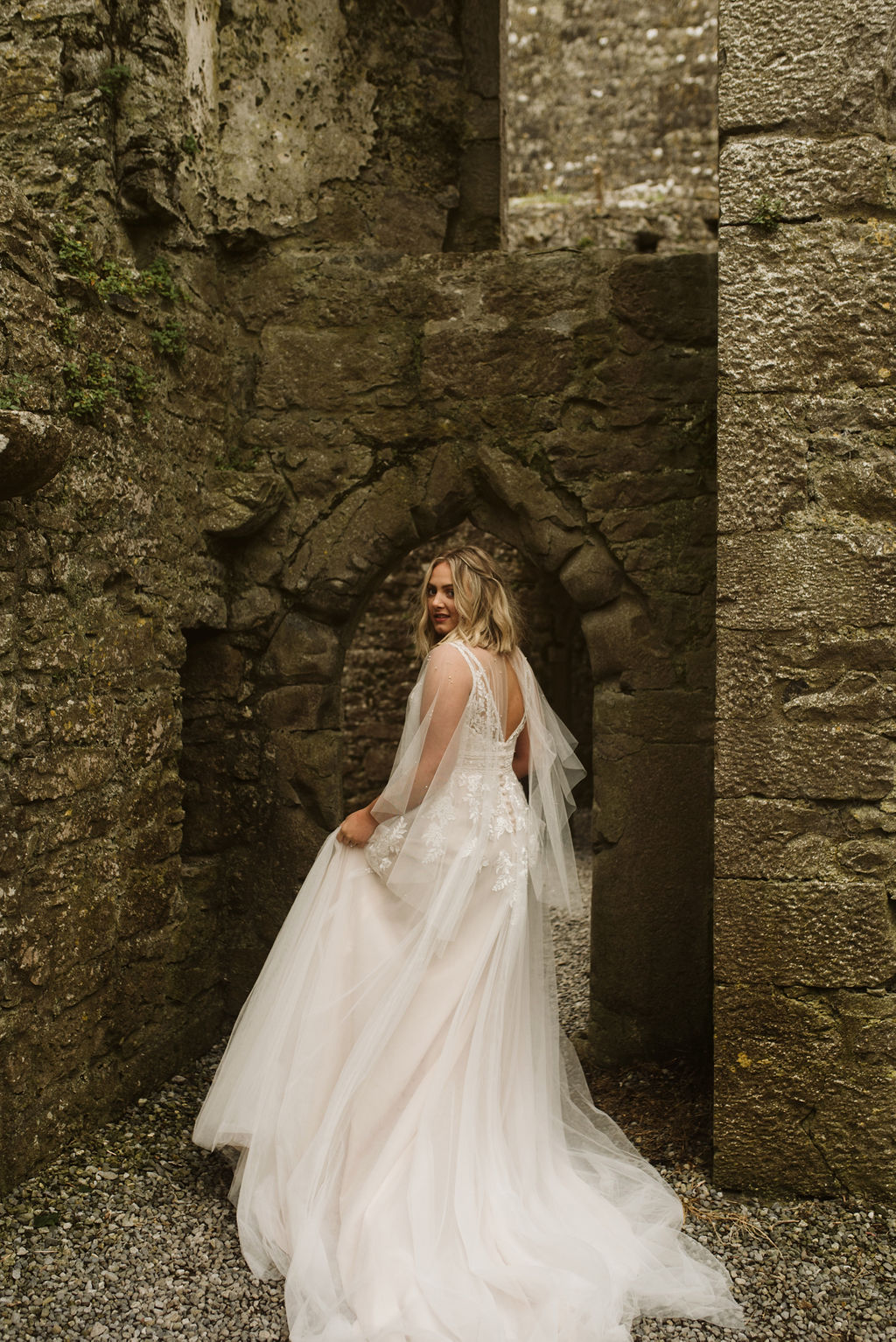 View of bride from the back looking over her shoulder wearing a lace and tulle aline wedding dress and tulle cape at Ross Errilly Friary in Ireland