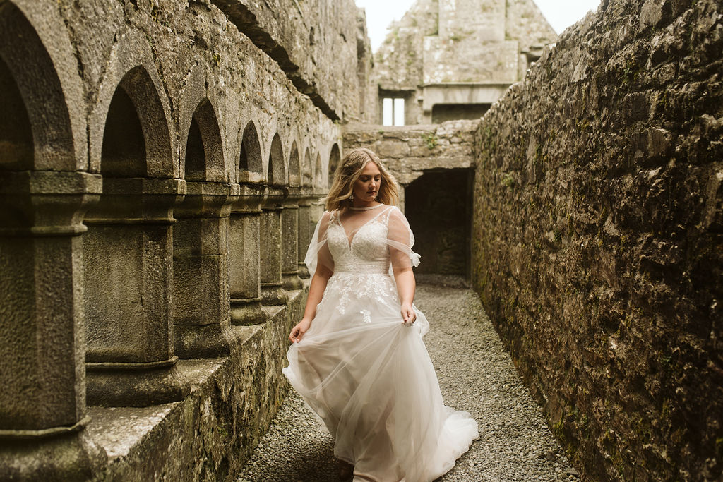Bride walking through Ross Errilly Friary in Ireland wearing a lace and tulle aline wedding dress with lace straps and a tulle cape