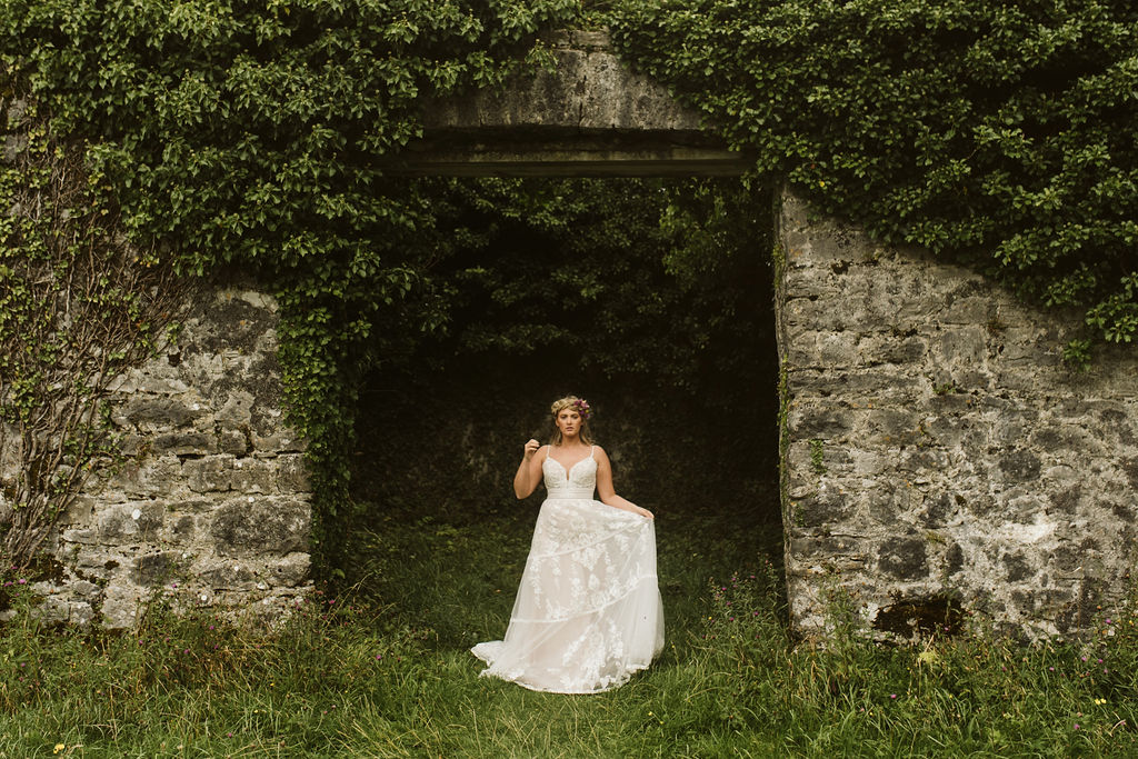 Bride wearing a lace boho wedding dress with straps stands in front of the abandoned Menlo Castle in Ireland