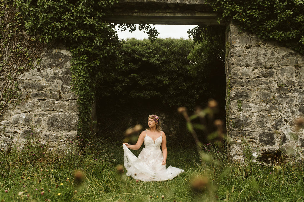 Bride kneels in the grass with flowers in her hair wears a lace ballgown wedding dress with waist detail and straps next to Menlo Castle in Ireland