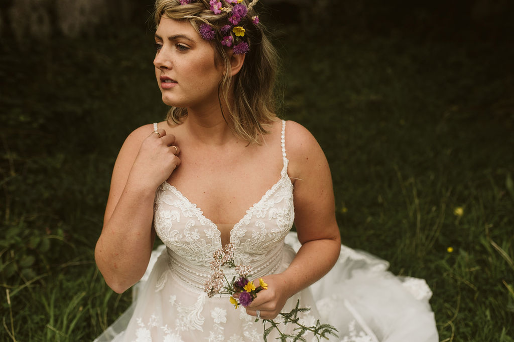 Bride with wildflowers in her braid kneels in the grass wearing a lace boho wedding dress with straps at Menlo Castle in Ireland