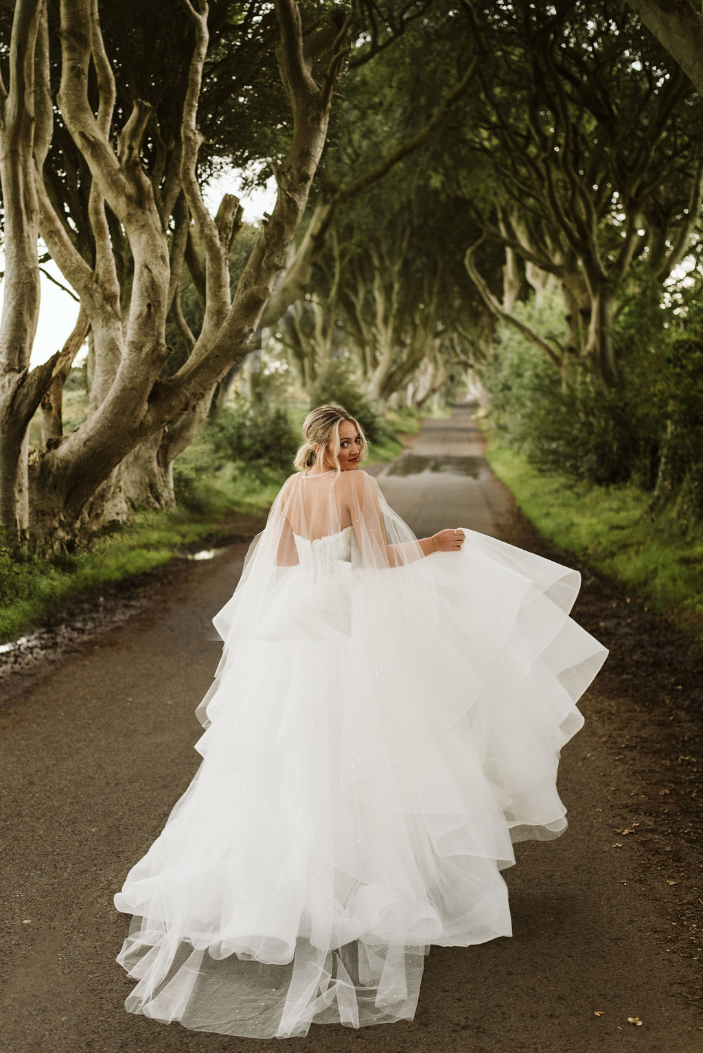 Bride looking over her shoulder in a lace and tulle horsehair ballgown wedding dress with a pearl and crystal tulle cape at The Dark Hedges in Northern Ireland
