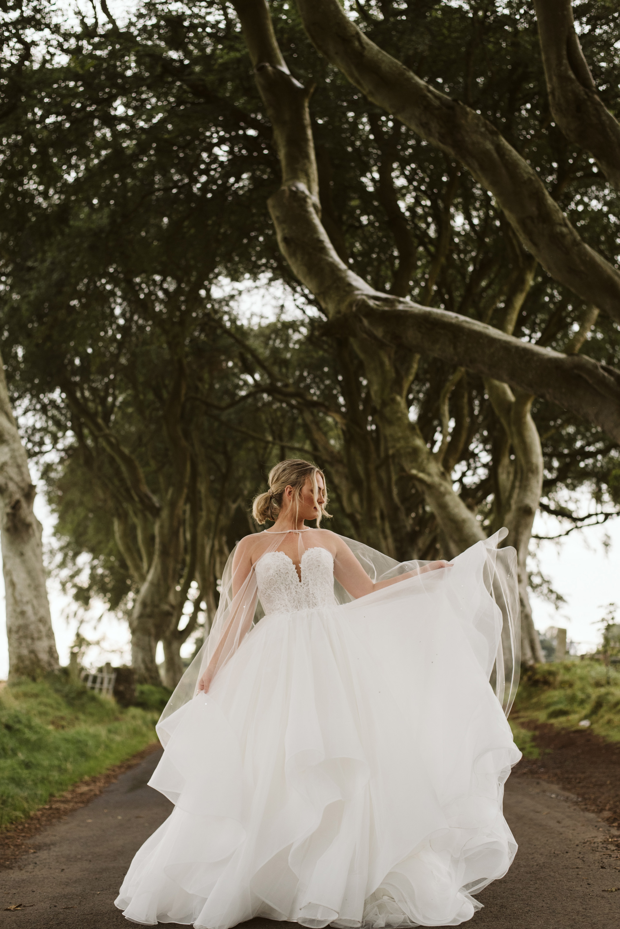 Bride plays with the skirt of her lace and tulle horsehair ballgown wedding dress while wearing a tulle cape at The Dark Hedges in Northern Ireland