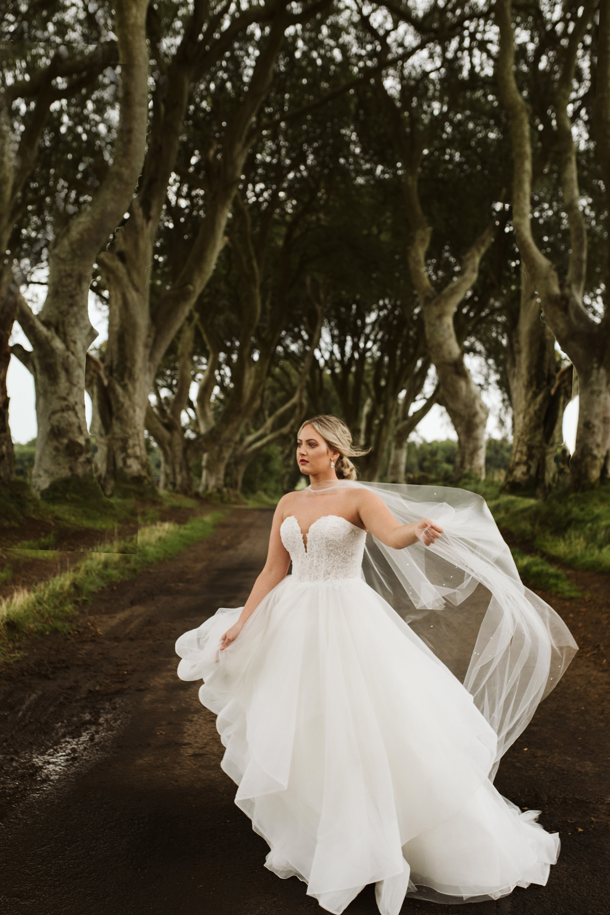 Bride wearing a lace and tulle horsehair ballgown wedding dress while her tulle cape blows in the wind at The Dark Hedges in Northern Ireland