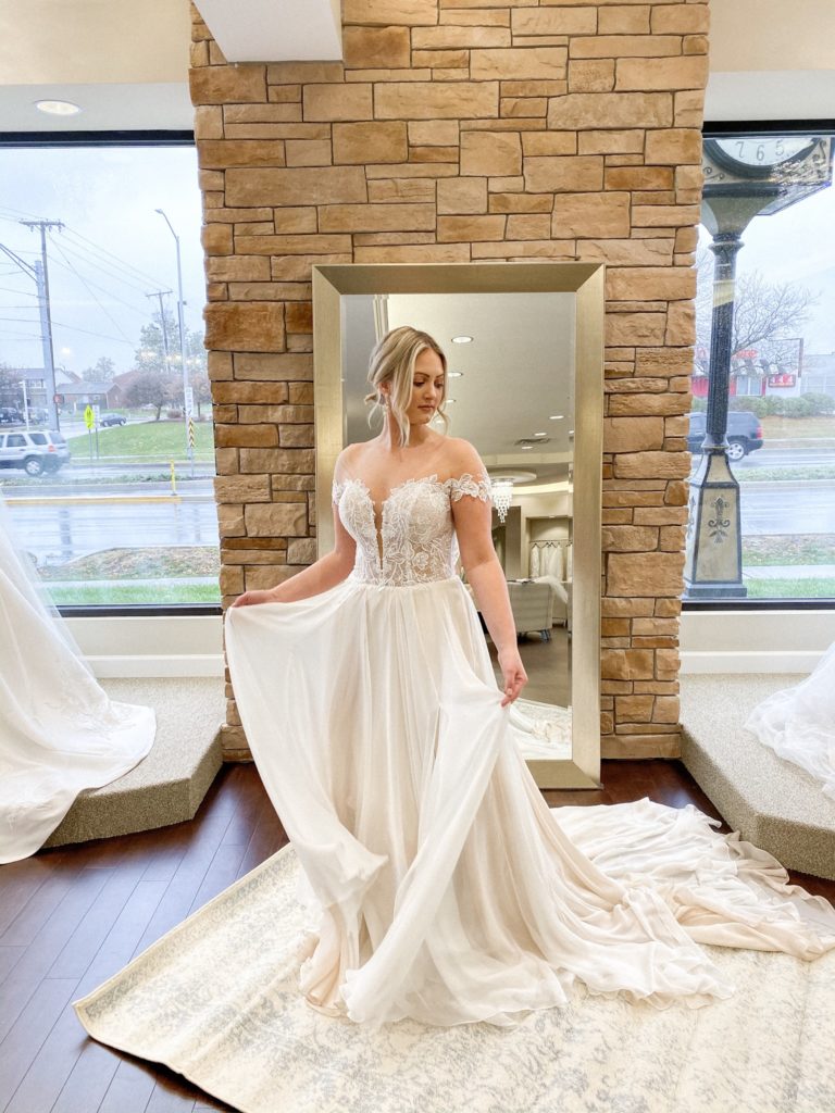 Bride stands in a chiffon skirt ballgown with off the shoulder sleeves at One Fine Day Bridal and Gown Boutique in Fort Wayne, Indiana